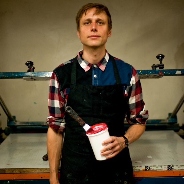 Zu sehen ist der Künstler Alex Hanke als Portrait in amerikanischer Perspektive. In der Hand hält er eine Dose mit roter Farbe und einem Spachtel darin steckend. Im Hintergrund ist eine Siebdruckmaschine zu sehen. Alex schaut leicht lächelnd in die Kamera.