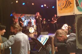 Colored Gigs Ausstellung mit Live-Musik Blick in die Ausstellungsatmosphäre mit Live-Band auf der Bühne. Im Vordergrund hängen einige Siebdruckposter von der Decke, Menschen sitzen, stehen und laufen im Vordergrund, wirken beschäftigt.