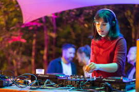 annalorenzana_F_ManuelLiebchen_3zu2 DJ Anna Lorenzana im roten Rollkragenpullover in Aktion. Hinter ihr ist Publikum und das Setting einer Freiluftveranstaltung mit Sonnensegel und Bäumen zu sehen. Die Szene ist effektvoll mit farbigem Licht ausgeleuchtet.
