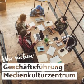 Zu sehen ist ein helles Bürozimmer mit Holzfußboden. Fünf Menschen sitzen mit Laptops, Klemmbrett, Notizbüchern, Tablet und Smartphones an einem Tisch und besprechen sich. Im Vordergrund ist folgende Schrift zu sehen: "Wir suchen: Geschäftsführung Medienkulturzentrum".