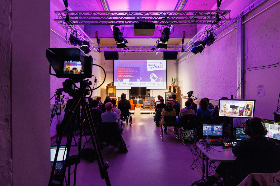 Bild von einer Veranstaltung mit magenta und violettem Licht, Bestuhlung, Teilnehmenden, Kamera und Bildschirmen