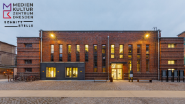 Zu sehen ist ein Gebäude aus rotem Ziegelstein mit Flachdach. Einige Fenster sind hell erleuchtet.