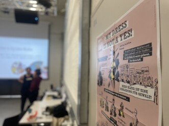 Blick in einen tiefen Raum, links unscharf im Hintergrund 2 Personen im Gespräch vor einem Beamerbild, rechts eine Wand mit Plakat.