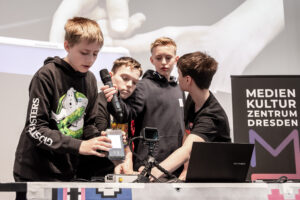 Vier Jugendliche stehen an einem Tisch mit technischem Equipment und Mikrofon vor einem Banner mit der Aufschrift 'MEDIEN KULTUR ZENTRUM DRESDEN'