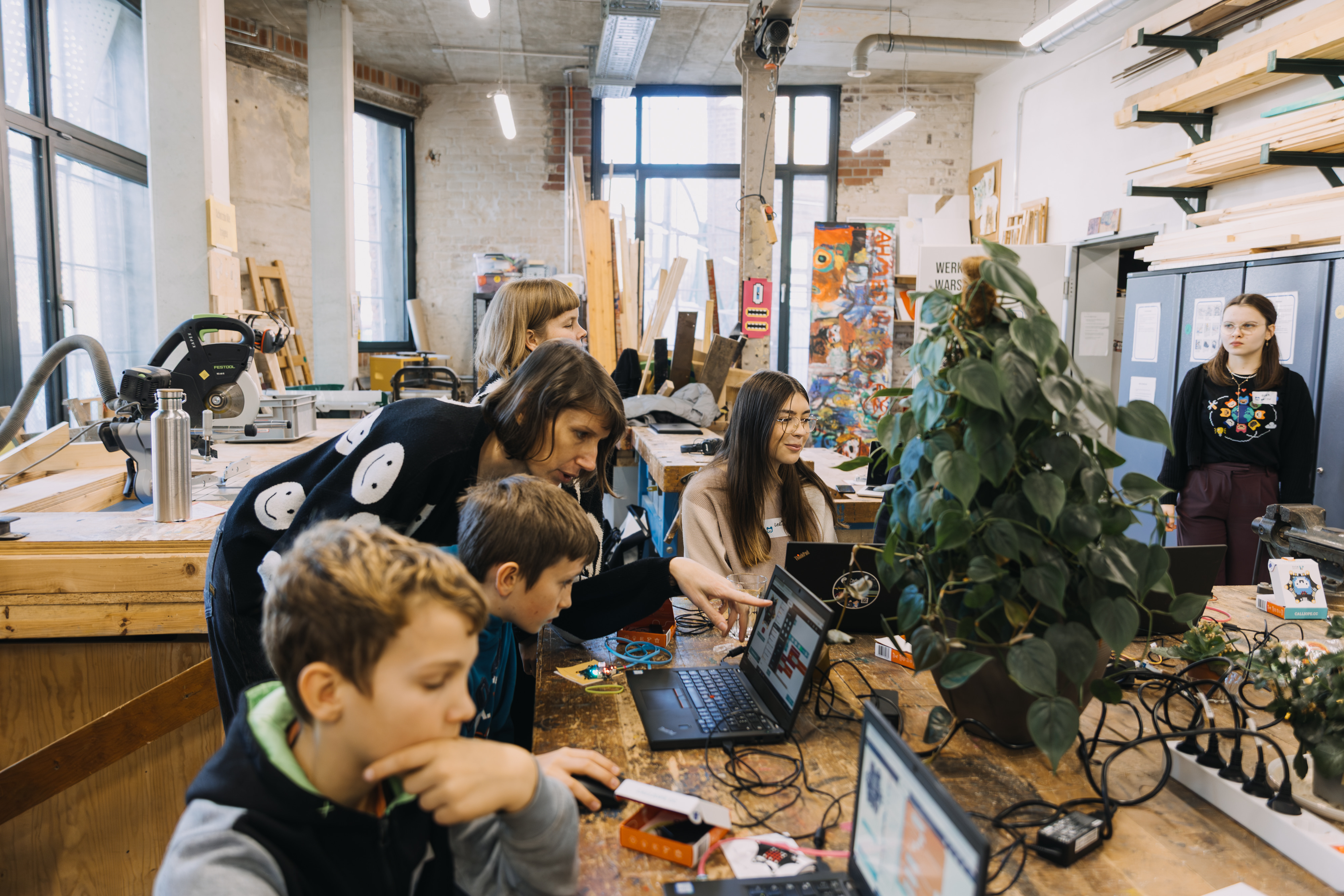 Jugendliche sitzen an einem langen Holztisch mit Laptops und elektronischen Bauteilen in einem hellen Werkraum, eine junge Frau steht vorne und spricht zu ihnen
