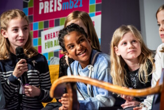 4 Kinder im Scheinwerferlicht auf einer Bühne, eins hält ein Mikrofon, alle lächeln, es scheint gute Stimmung zu sein.