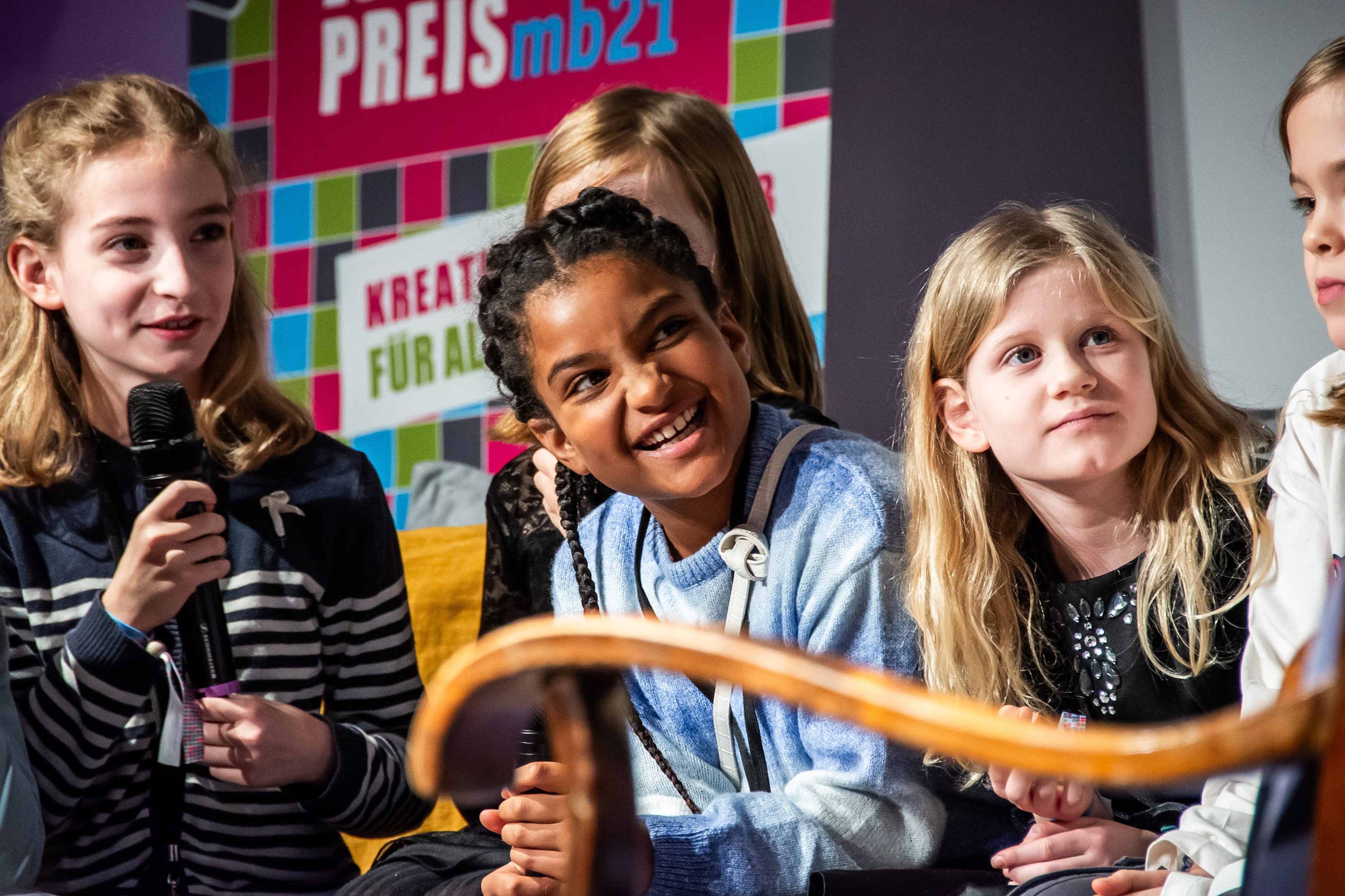 4 Kinder im Scheinwerferlicht auf einer Bühne, eins hält ein Mikrofon, alle lächeln, es scheint gute Stimmung zu sein.