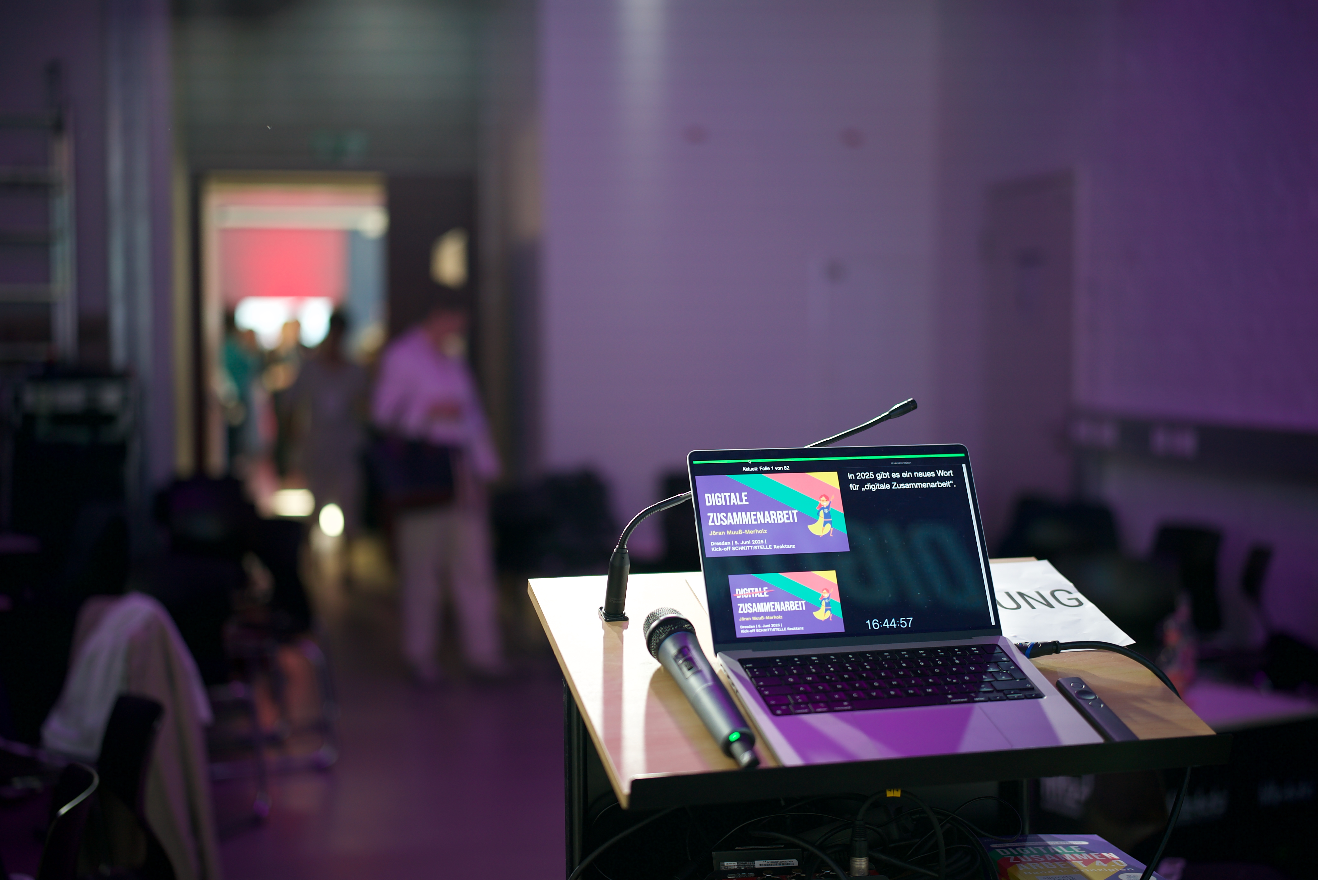Laptop mit Präsentationsfolien und Mikrofon auf einem Pult in einem dunklen Raum mit unscharfen Personen im Hintergrund