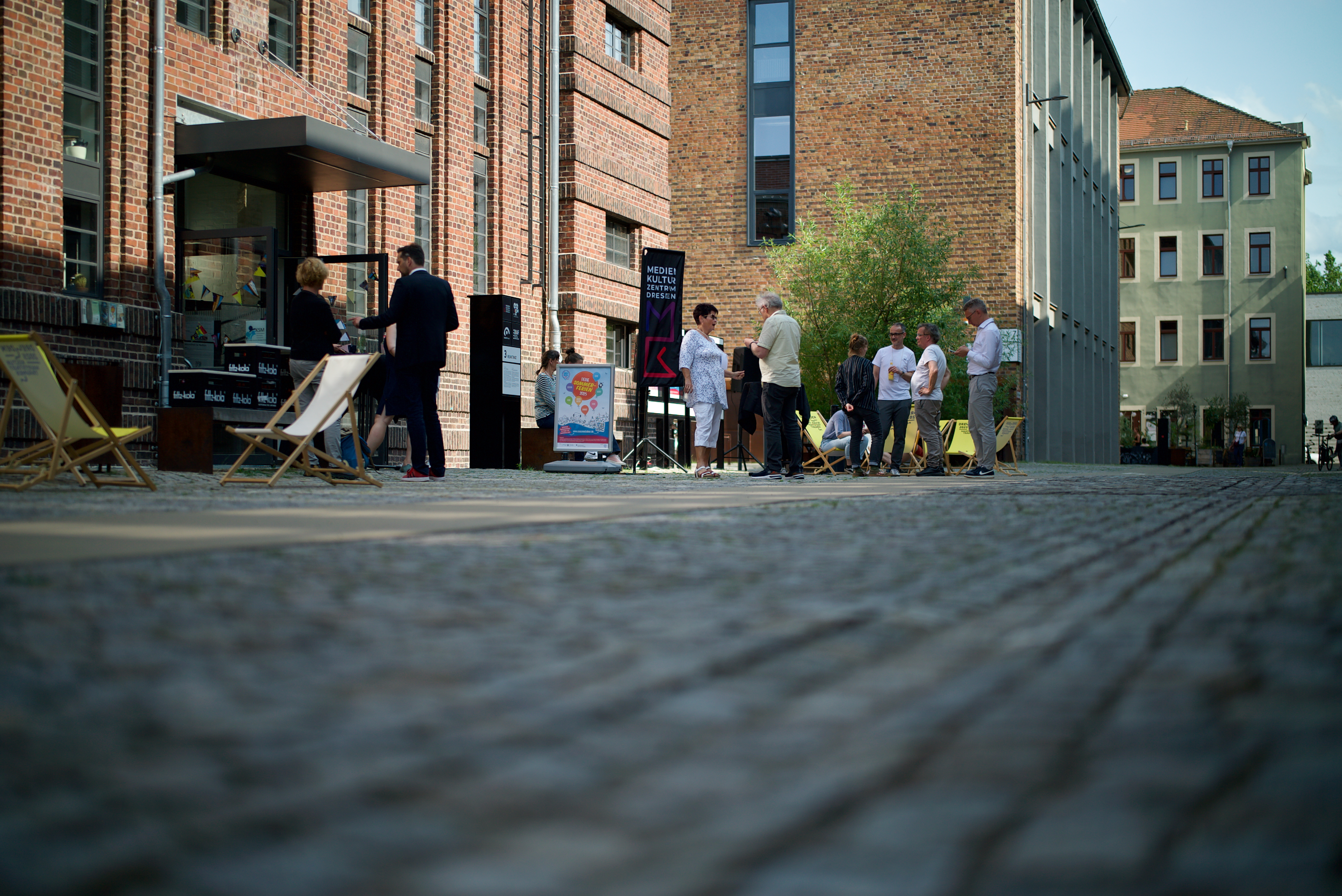 Menschen stehen und unterhalten sich vor einem Backsteingebäude auf einem gepflasterten Platz bei Tageslicht
