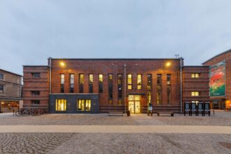 Ansicht eines Backsteingebäudes mit Flachdach in der Abenddämmerung mit erleuchteten Fenstern.