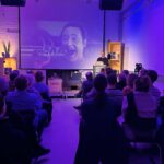 Blick in einen Raum voller Publikum mit athmosphärischer Wandbeleuchtung in Blau und Violett. Im Hintergrund eine Bühne mit einer Präsentation und einer vortragenden Person.