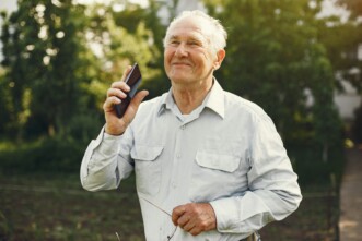 Älterer Mann in der Natur mit Smartphone und Brille in der Hand, glücklich lächelnd