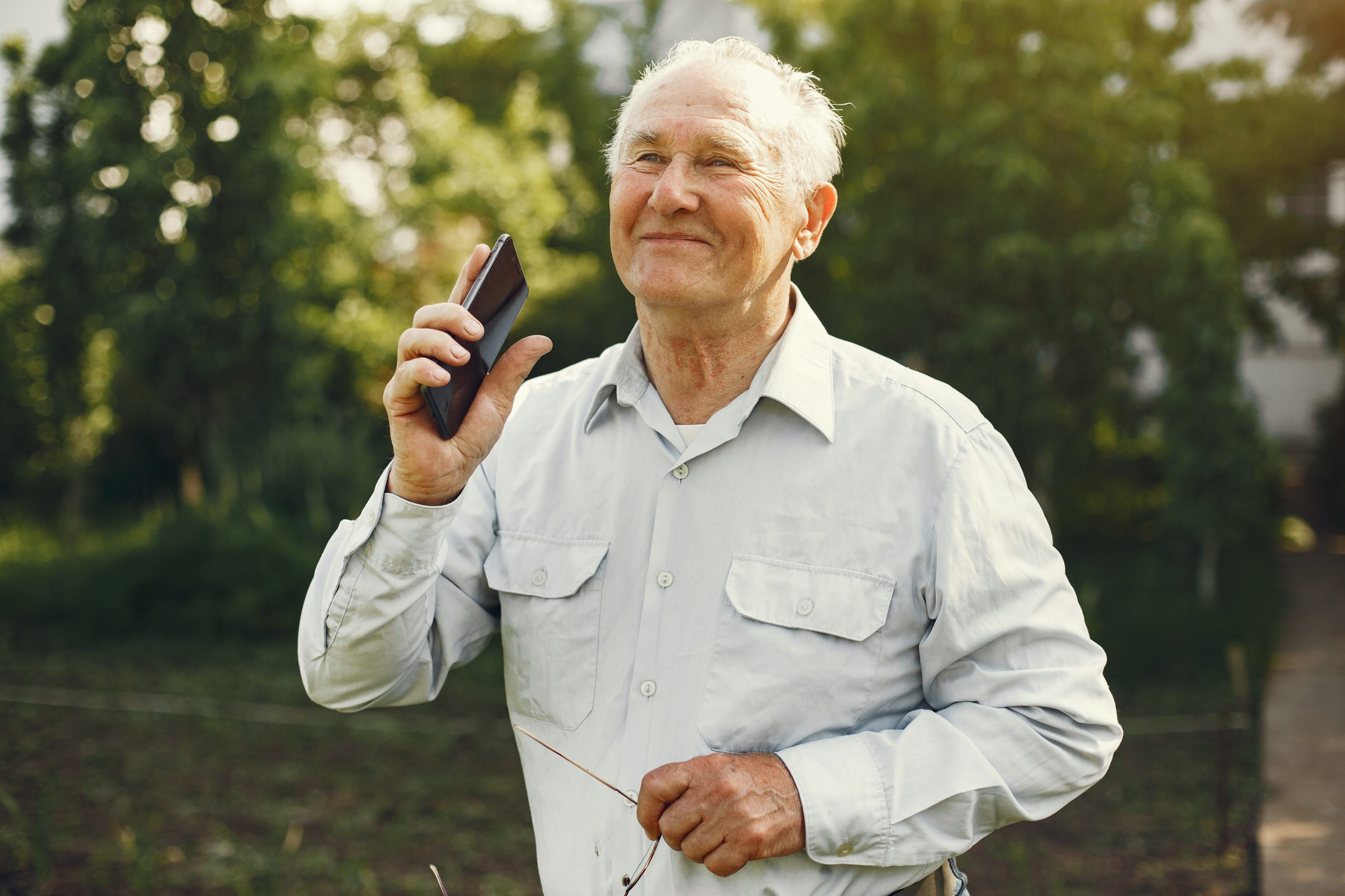 Älterer Mann in der Natur mit Smartphone und Brille in der Hand, glücklich lächelnd