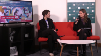 Still aus dem Videointerview mit Kristina Richter und Lene Schargitz. Zu sehen sind die beiden in einer Gesprächssituation mit Mikrofon auf einem roten Sofa.
