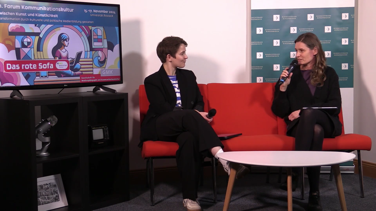 Still aus dem Videointerview mit Kristina Richter und Lene Schargitz. Zu sehen sind die beiden in einer Gesprächssituation mit Mikrofon auf einem roten Sofa.