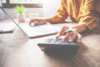 Ein Schreibtisch mit Laptop und einem Taschenrechner und zwei Arme einer Frau in einer gelben Bluse, eine Hand auf der Laptoptastatur, eine Hand mit einem Stift zwischen den Fingern am Taschenrechner. Warmes Licht und eine kleine Pflanze auf dem Holztisch.