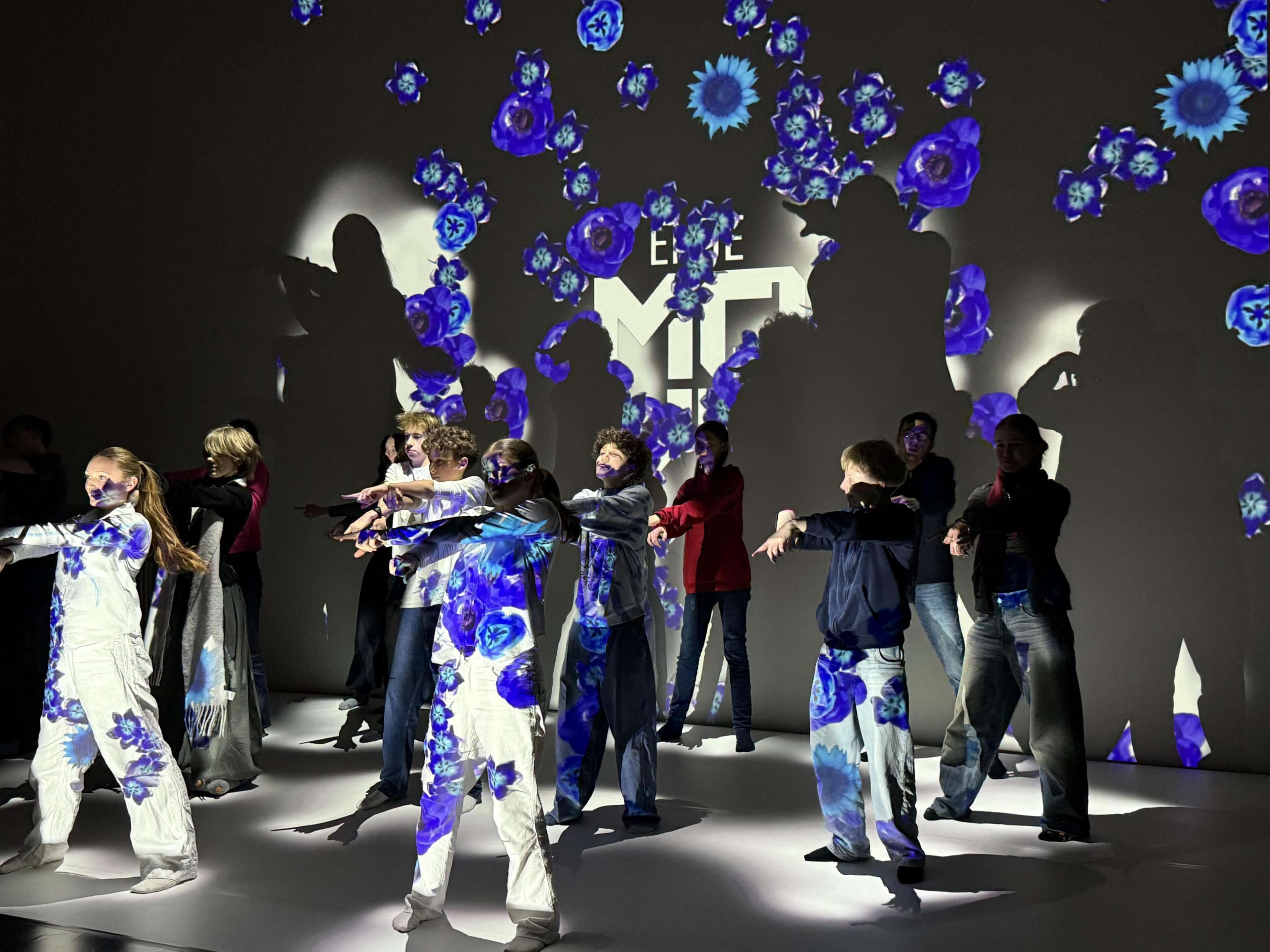 Gruppe von Jugendlichen, einige in weißen Overalls mit blauen Blumenmustern tanzt vor einer Wand mit projizierten blauen Blumen und Schatten