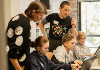 drei Jugendliche am Laptop, dahinter 2 Frauen, die darauf schauen, was die Jugendlichen machen