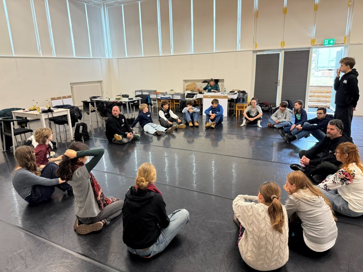 Gruppe von Menschen sitzt im Kreis auf dem Boden eines großen Raums mit Tischen und Stühlen im Hintergrund
