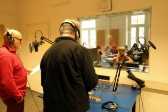 Blick in das Hörfunkstudio. Zwei Senioren stehen in der Sprecherkabine, sie haben Kopfhörer auf und stehen vor Mikrofonen. Sie sind von hinten bzw. von der Seite zu sehen. Durch die Scheibe blickt man in den Moderationsraum. Dort sitzen weitere Senioren, sie sind von vorne zu sehen.
