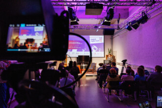 Blick durch eine Kamera auf einen Vortrag mit Publikum in einem Veranstaltungsraum mit Projektionsleinwand und Beleuchtungstechnik