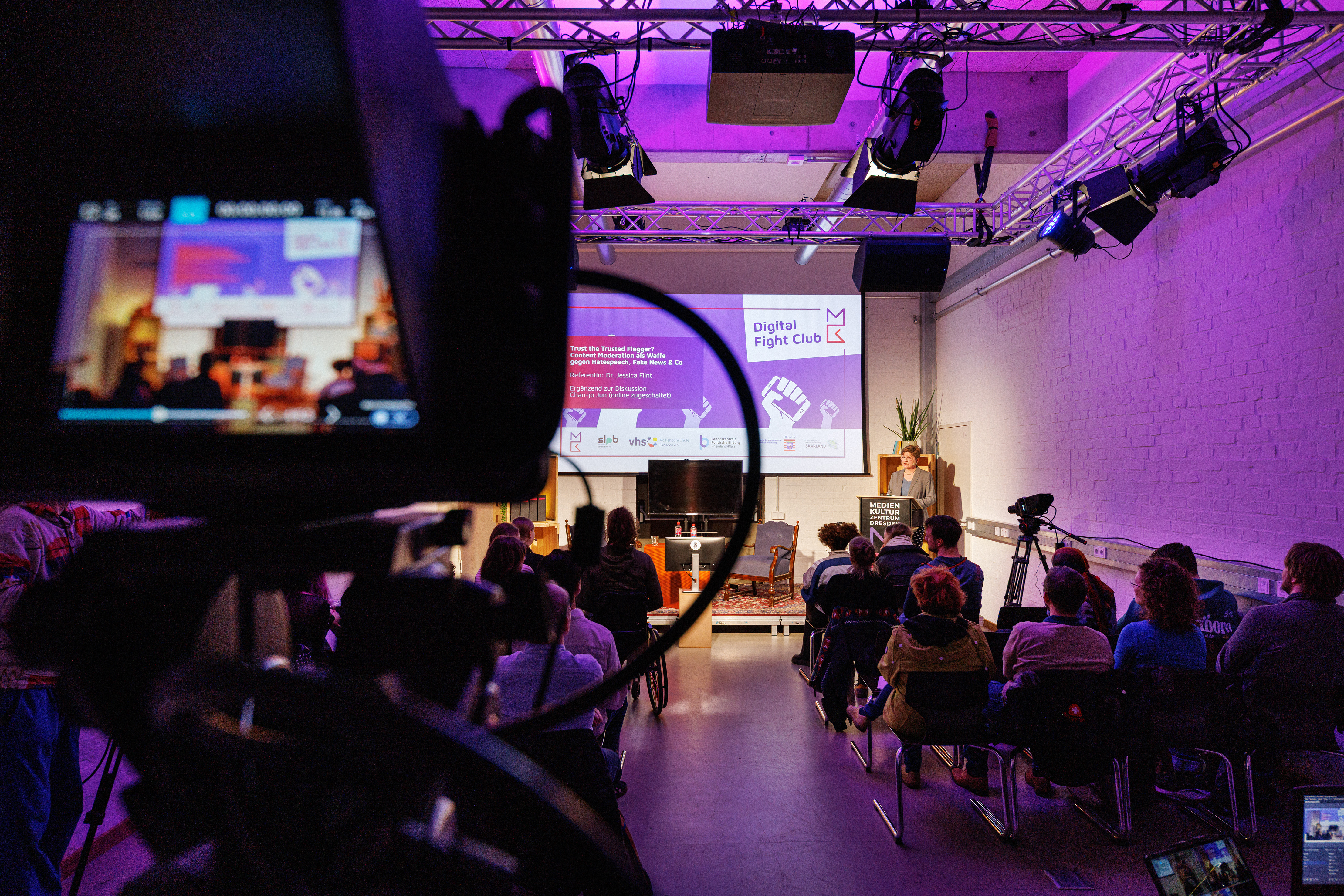 Blick durch eine Kamera auf einen Vortrag mit Publikum in einem Veranstaltungsraum mit Projektionsleinwand und Beleuchtungstechnik