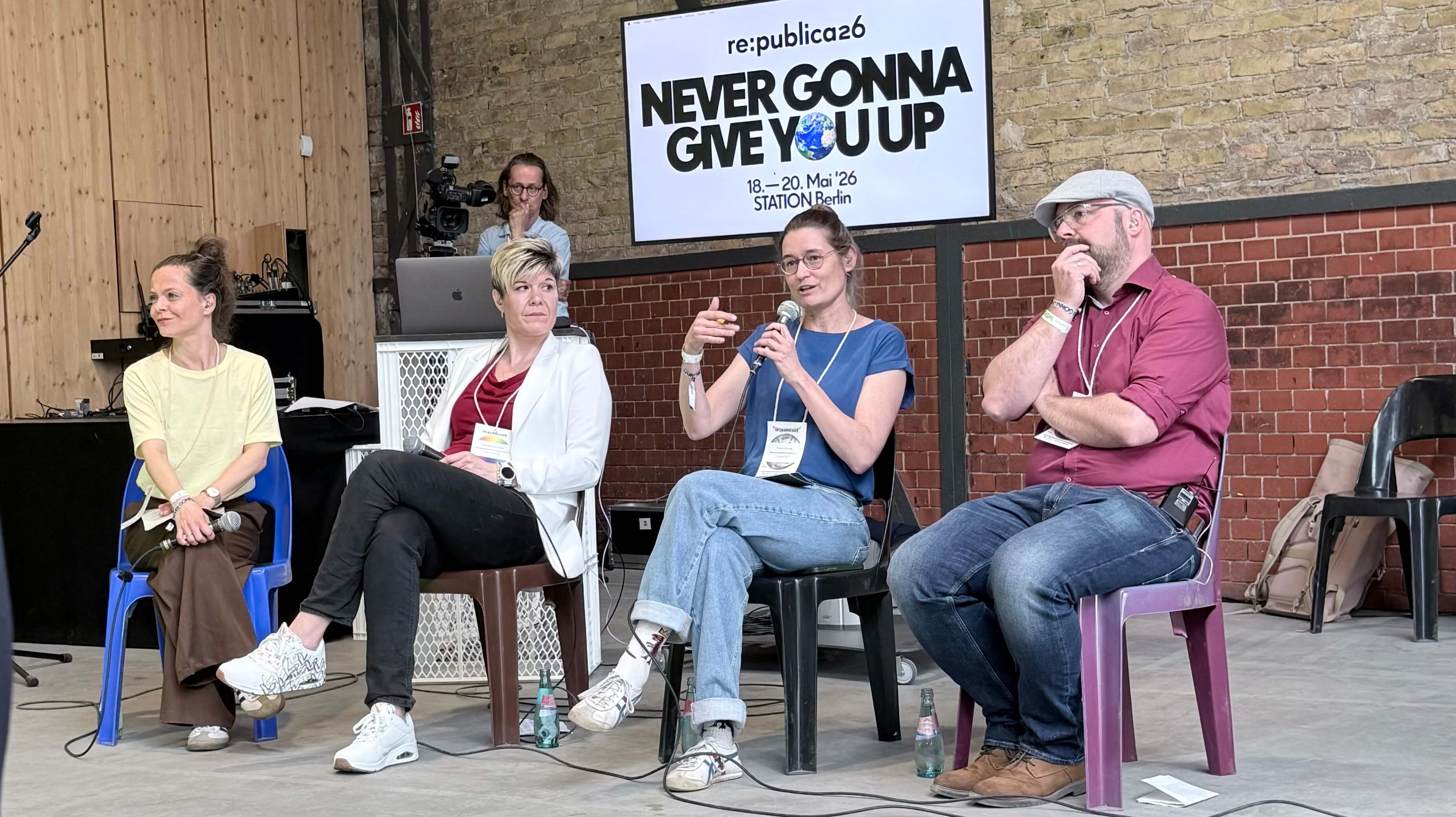 Vier Personen sitzen auf Stühlen vor einer Wand mit Holz- und Backsteinelementen, im Hintergrund ein Bildschirm mit dem Text 'NEVER GONNA GIVE YOU UP'