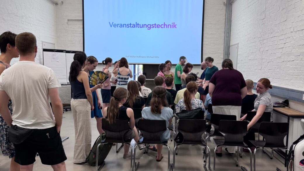 Gruppe von Menschen in einem Raum mit weißer Backsteinwand vor einer Leinwand mit dem Text 'Veranstaltungstechnik'