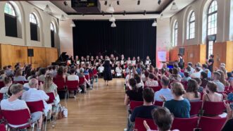 Großer Saal mit Publikum auf roten Stühlen und Chor auf Bühne vor schwarzem Vorhang
