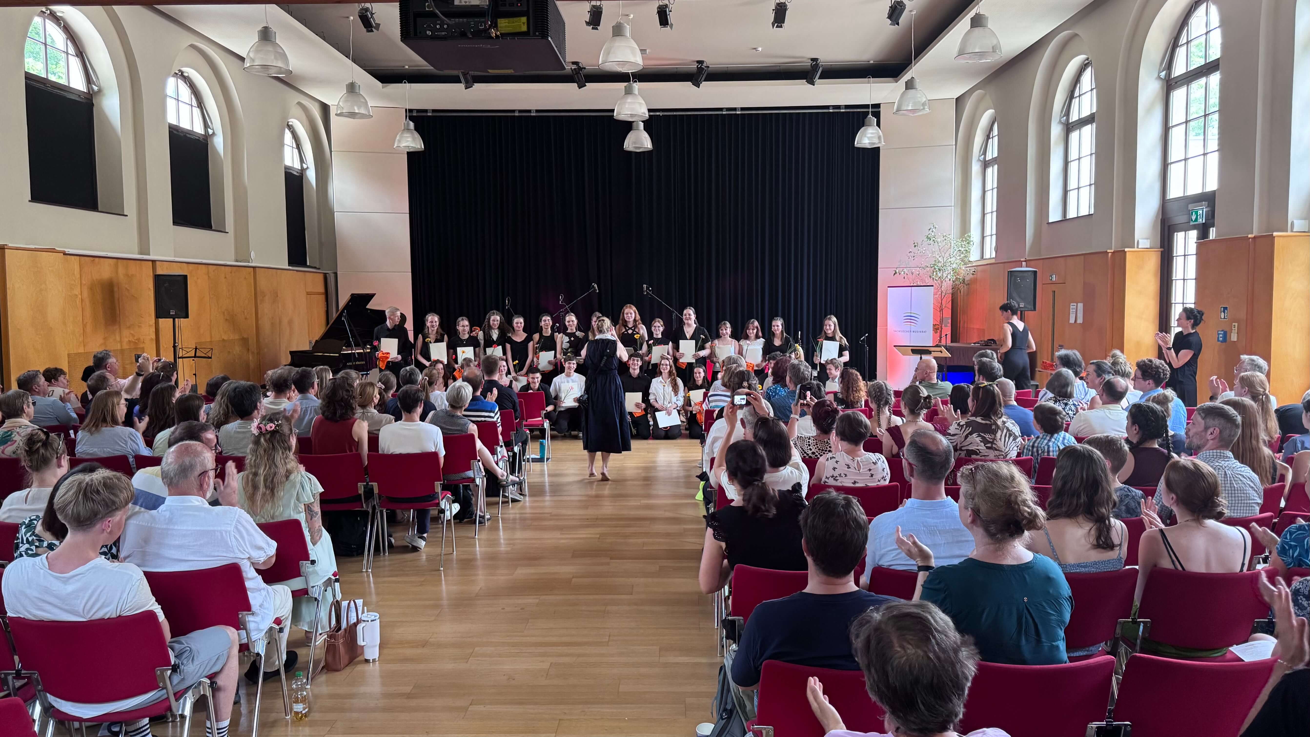 Großer Saal mit Publikum auf roten Stühlen und Chor auf Bühne vor schwarzem Vorhang