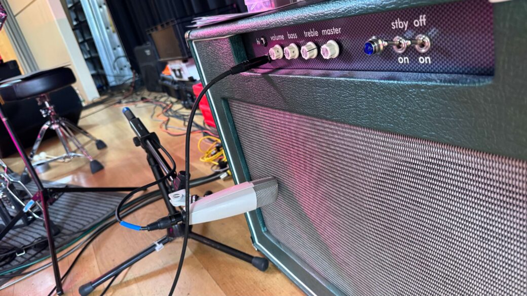 Nahaufnahme eines Gitarrenverstärkers mit angeschlossenem Mikrofon in einem Raum mit Musikinstrumenten und Mikrofonstativen im Hintergrund