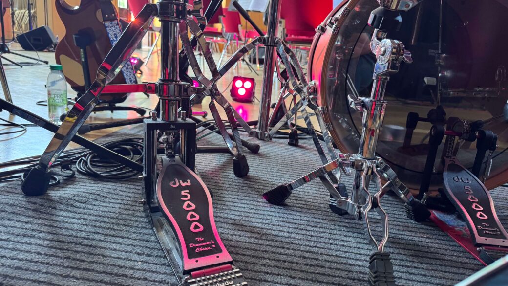 Nahaufnahme von Schlagzeugpedalen und Ständern auf einem Teppichboden mit Gitarren im Hintergrund